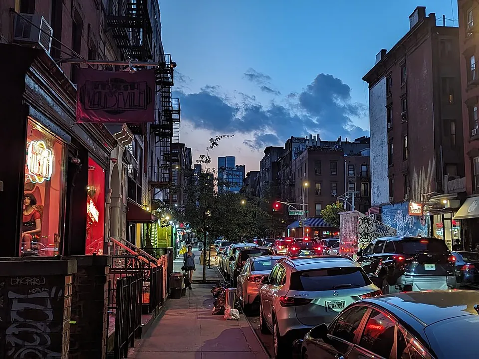 An East Village street in Manhattan at dusk, a neon-lit shop window beside a line of parked cars